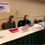Two women sitting at outreach table labeled "Voting Information - LWV", helping a visitor
