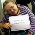 Woman sitting in chair holding sign that says "Disability issues matter. I Vote."